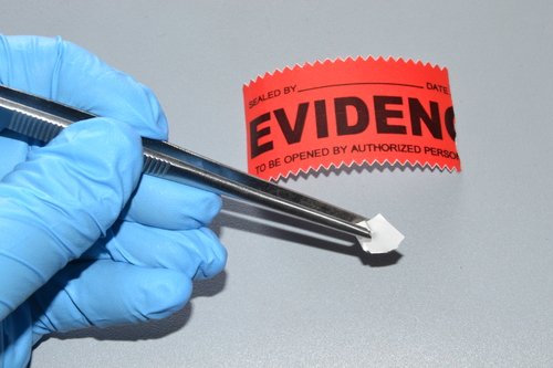 Expert witness wearing blue surgical gloves examining evidence with tweezers and red label.